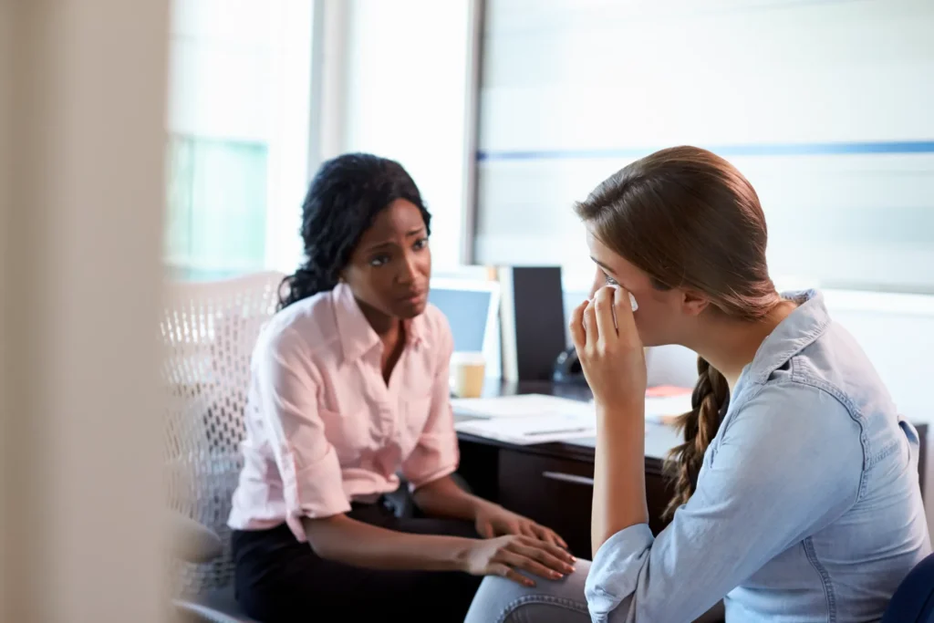 Foto de uma colaboradora visivelmente abalada emocionalmente, buscando apoio psicológico com uma profissional da área durante o expediente, mostrando a importância da discussão de riscos psicossociais no ambiente de trabalho e seu impacto na produtividade e bem-estar corporativo.