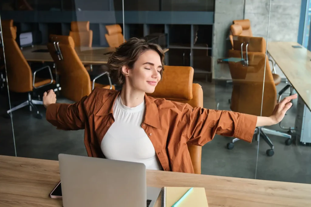 Foto de mulher jovem sorrindo, relaxada no escritório, praticando alongamento e tendo um momento de bem-estar durante o dia de trabalho.