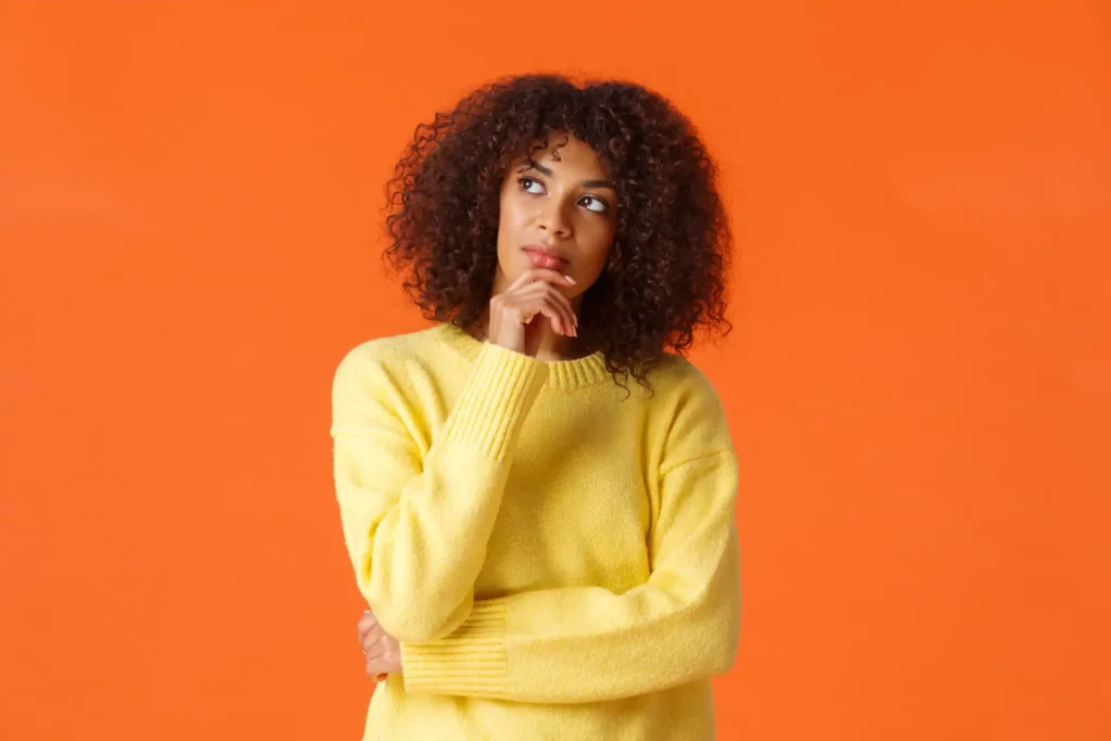 Foto de uma mulher negra pensativa, vestindo suéter amarelo, em um fundo laranja, representando considerações importantes de pacientes antes de agendar um psicólogo.