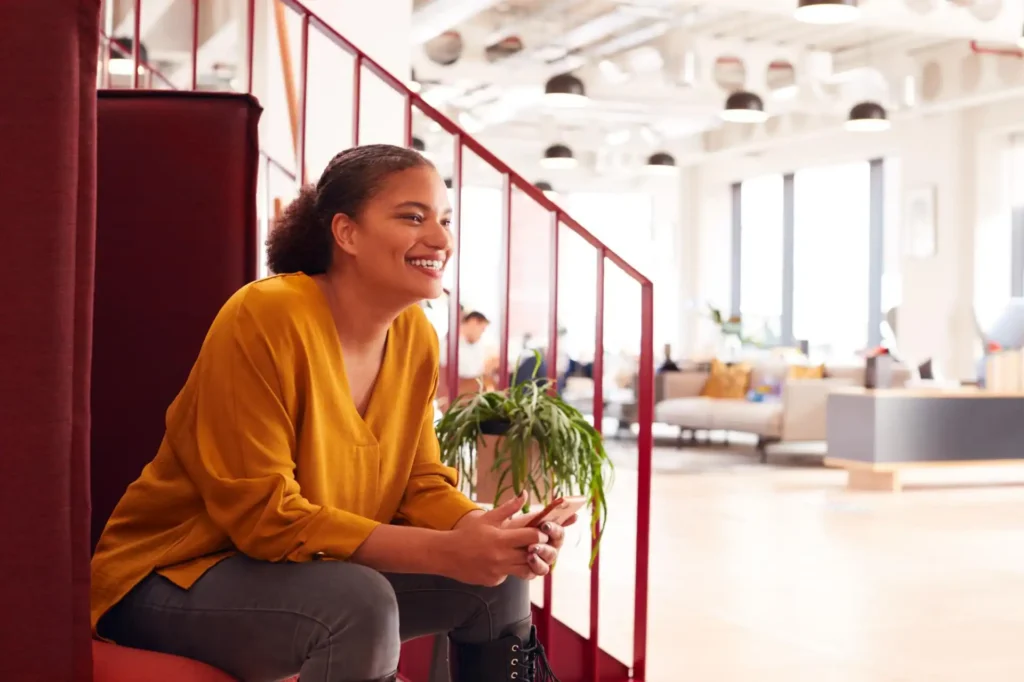 Foto de uma colaboradora em espaço corporativo moderno expressando alegria e leveza, mostrando a importância do espaço zen para a saúde mental no trabalho, ajudando nos níveis de produtividade e engajamento entre equipes.